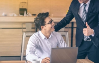 An employee on a laptop smiles at his supervisor as he gives him praise