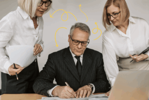 A businessman writing as two coworkers advise him. Squiggly arrows surround his head, signifying thoughts and ideas.
