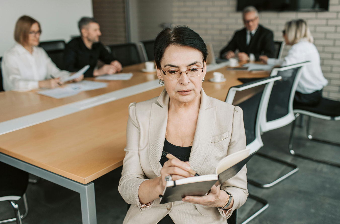 A businesswoman standing in front of a table, her team convening at the table behind her.