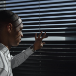 A man peering through the blinds at a window.